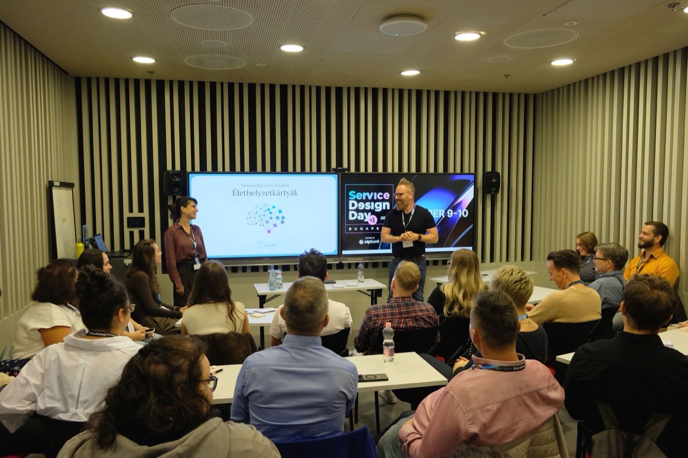 Konferenciaterem a Service Design Day-en: mintegy huszonöt résztvevő ül asztaloknál, elöl két előadó. A kivetítőn az Élethelyzetkártyák címlapja, mellette a Service Design Day arculata.